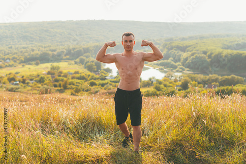 A photo of a man showing his muscles on a hill looking at the camera on a sunny day .