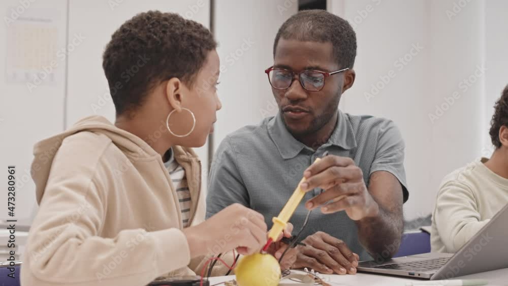 Waist-up of young Black teacher and female secondary school student sitting at table in classroom, doing experiment with electricity and lemon, talking