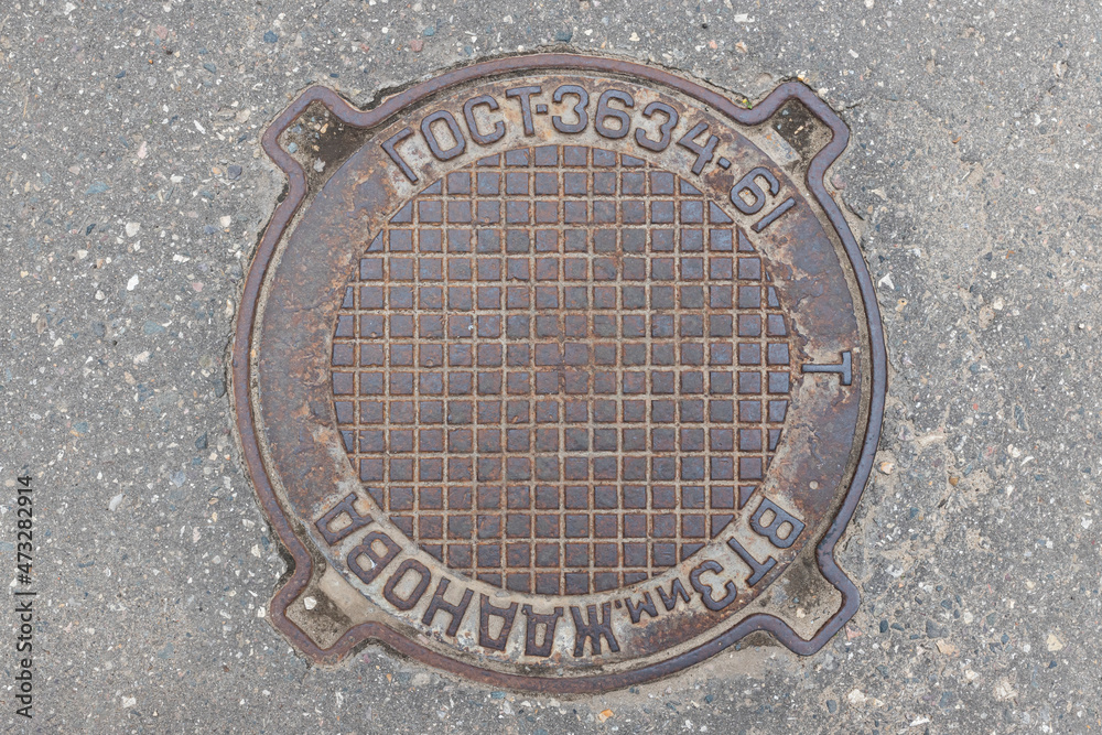 Old iron manhole cover on the city street. Translation: abbreviation for State Standard, Zhdanov Plant