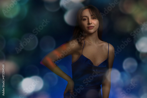 Young and cute brunette woman in blu dress posing on dark blue background with christmas party lighting bokeh. Free space for text