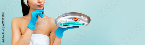 Banner of cosmetology. Close up portrait of a thoughtful young woman in medical gloves choosing syringes from medical kidney dish. Blue background. Copy space. Concept of plastic surgery