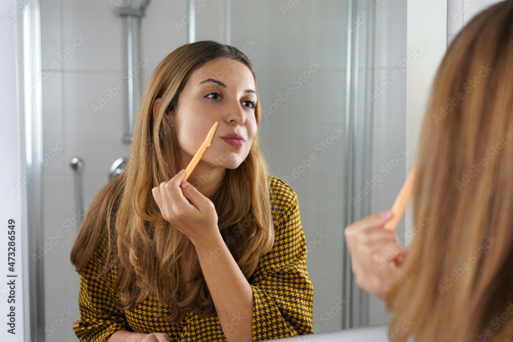 Facial hair removal. Beautiful young woman shaving her face by razor at  home. Pretty woman using shaver on bathroom. Stock 写真 | Adobe Stock
