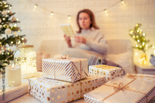 Woman sitting on couch near christmas tree and packing handmade gift boxes putting it on the table. Presents for family.
