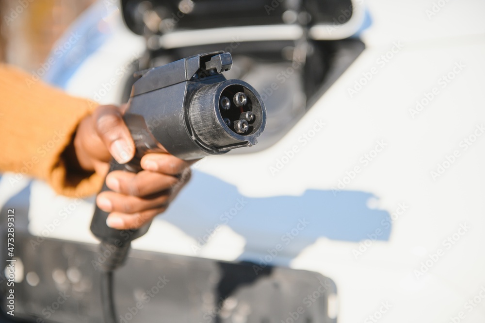 Fototapeta premium Close up of male hand holding electric charging plug for luxury red car. African businessman charging his auto on station. Eco friendly transport.
