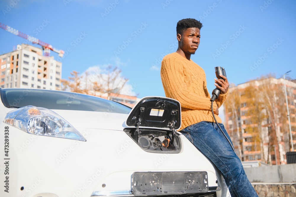 Urban, electric vehicle, eco concept. Young black skinned man, waiting for his electric car charging Urban, electric vehicle, eco concept. Young black skinned man, waiting for his electric car charging