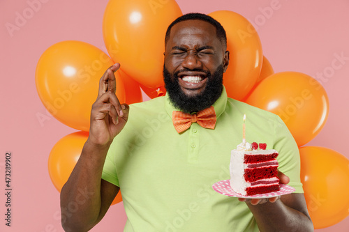 Young black gay man 20s wear green t-shirt bow tie hold bunch of air inflated helium balloons celebrate birthday party sweet cake with candle cross fingers isolated on plain pastel pink background © ViDi Studio