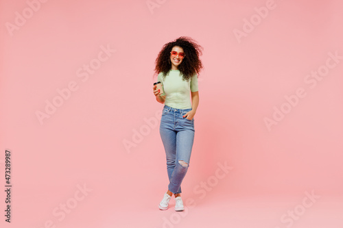Full size body length happy young curly latin woman 20s wears casual clothes hold takeaway delivery craft paper brown cup coffee to go isolated on plain pastel light pink background studio portrait.