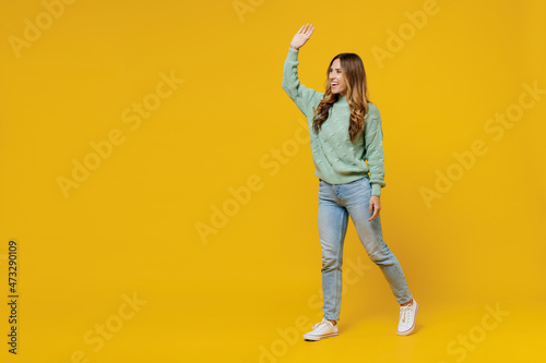 Full body young smiling satisfied happy fun woman 30s wearing green knitted sweater walk go strolling waving hand isolated on plain yellow color background studio portrait. People lifestyle concept. © ViDi Studio