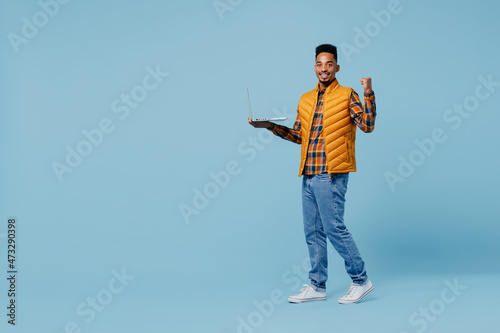 Full size body length young black man 20s years old wear yellow waistcoat shirt hold use work on laptop pc computer doing winner gesture isolated on plain pastel light blue background studio portrait