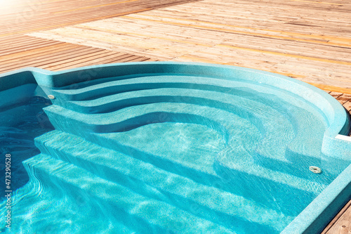 New modern fiberglass plastic swimming pool entrance step with clean fresh refreshing blue water on bright hot summer day at yard or resort hotel spa area. Wooden flooring deck of teak or larch board photo