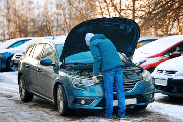 Man stands under open hood of modern car, checking engine oil level in winter season on parking lot. Man check fluid levels in winter. Winter car care