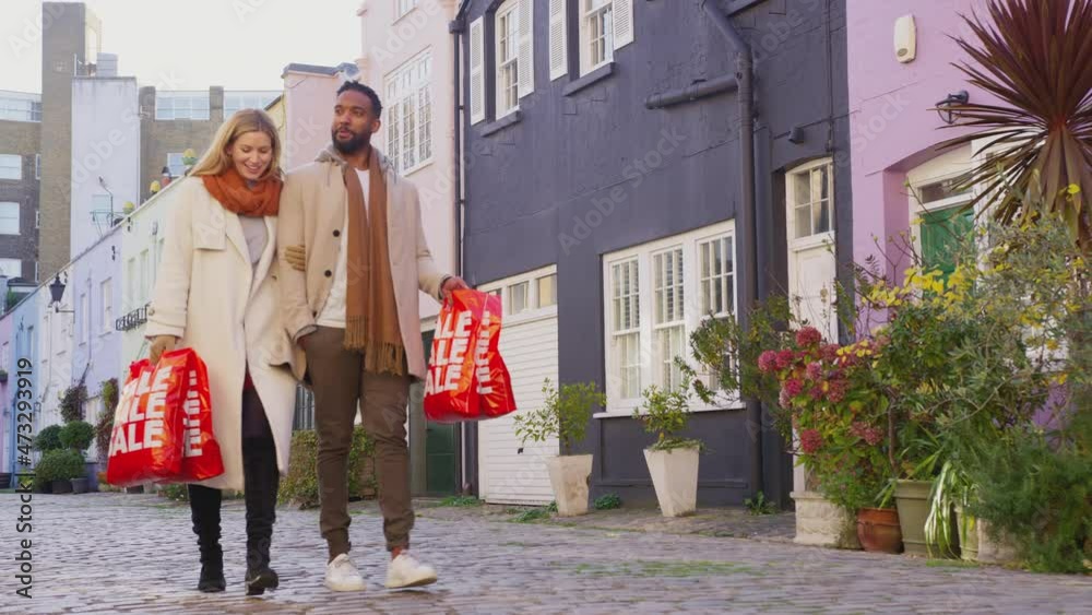 Multi-cultural couple arm in arm as they walk along cobbled mews street on visit to city in autumn or winter carrying sale shopping bags - shot in slow motion Multi-cultural couple arm in arm as they walk along cobbled mews street on visit to city in autumn or winter carrying sale shopping bags - shot in slow motion