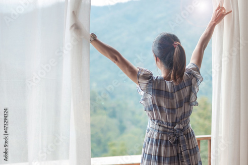 happy woman stretching near window, young adult female rising arms and looking mountain view in the morning. relaxing and lifestyle concepts