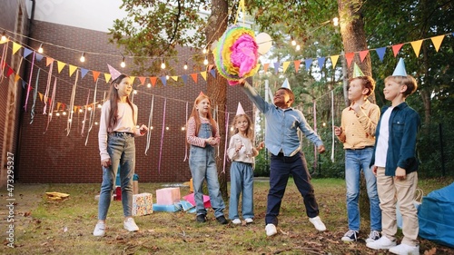 Group of kids smashing with a bat colorful pinata in the spring field at the park with balloons. Children playing outdoor with happy emotions photo