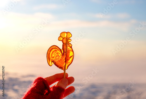 Sweet lollipop cockerel on stick in the hand on sky and snow background