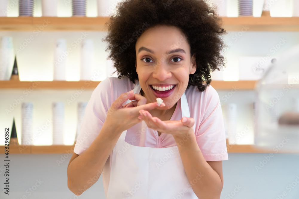 A beautiful mixed-race woman is smiling behind the stand of an ice cream parlor and trying the products. A beautiful mixed-race woman is smiling behind the stand of an ice cream parlor and trying the products.