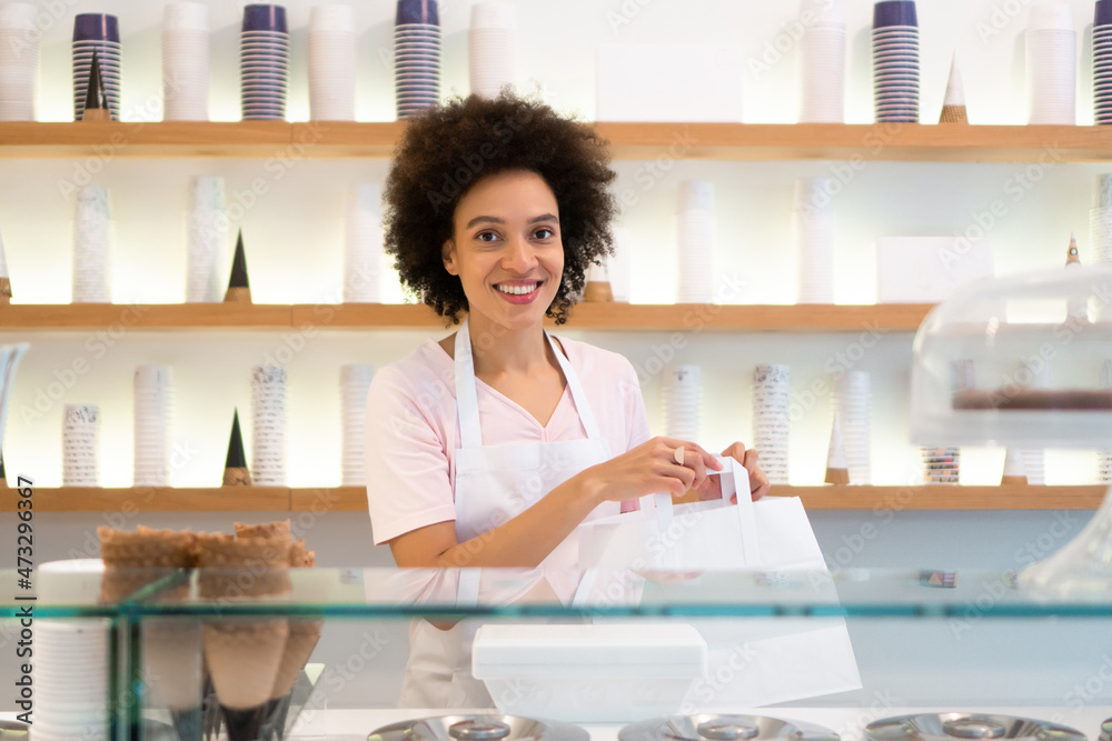 A beautiful mixed-race woman is smiling behind the stand of an ice cream parlor and giving the takeaway order in a paper bag. A beautiful mixed-race woman is smiling behind the stand of an ice cream parlor and giving the takeaway order in a paper bag.