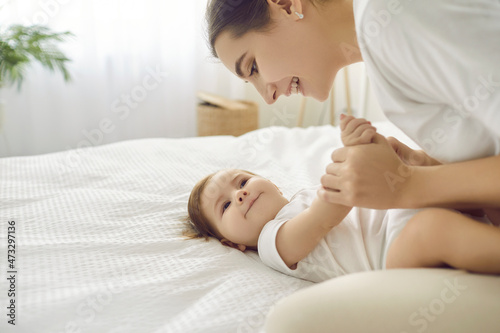 Happy mother playing with her baby. Charming infant looking at mom with cute, funny face expression. Beautiful young mommy and her little child cuddling on clean white bed in nursery room or bedroom