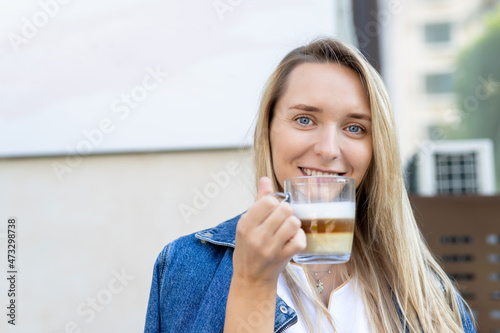 Portrait of young adult attractive happy woman enjoy smiling and hold cup drinking hot cappuccino coffee against street cafe wall in morning outdoors. Pretty female person wera jeans jacket outside photo