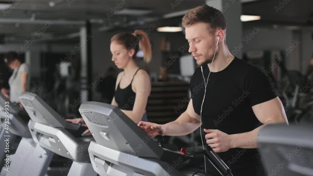 A young athletic man is running on a treadmill. He is putting on headphones and listening to music. A woman is running next to him. They're in the gym. 4K A young athletic man is running on a treadmill. He is putting on headphones and listening to music. A woman is running next to him. They're in the gym. 4K