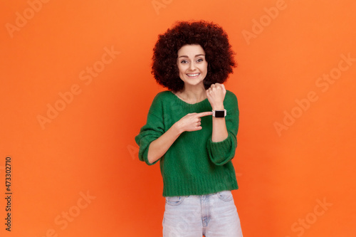 Good looking positive woman with Afro hairstyle wearing green casual sweater pointing at wristwatch on hand and smiling, showing clock, happy hours. Indoor studio shot isolated on orange background.