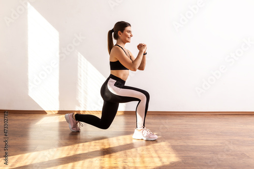 Slim female doing sport lunge exercise  standing one knee  warms up training muscles  butts workout  wearing black sports top and tights. Full length studio shot illuminated by sunlight from window.