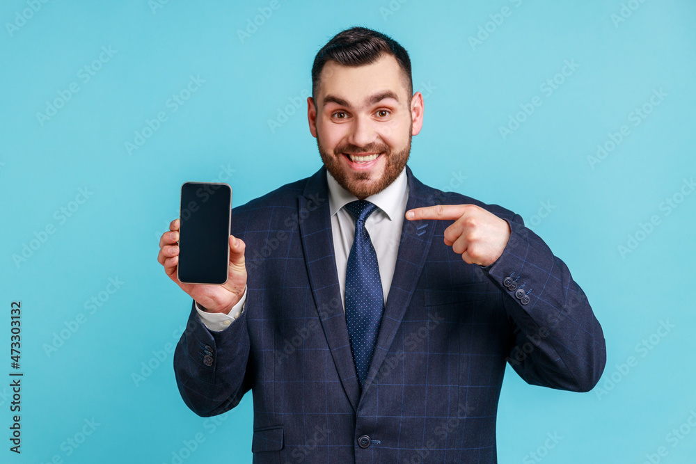 Happy cheerful beaded man in dark suit pointing finger at smartphone with empty screen looking at camera with toothy smile, freespace for adv. Indoor studio shot isolated on blue background. Happy cheerful beaded man in dark suit pointing finger at smartphone with empty screen looking at camera with toothy smile, freespace for adv. Indoor studio shot isolated on blue background.