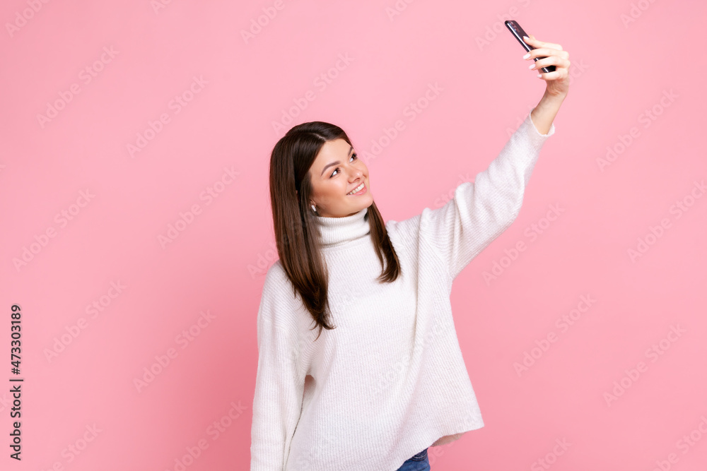 Brunette young female taking selfie or broadcasting livestream, looking smiling at cell phone camera, wearing white casual style sweater. Indoor studio shot isolated on pink background. Brunette young female taking selfie or broadcasting livestream, looking smiling at cell phone camera, wearing white casual style sweater. Indoor studio shot isolated on pink background.