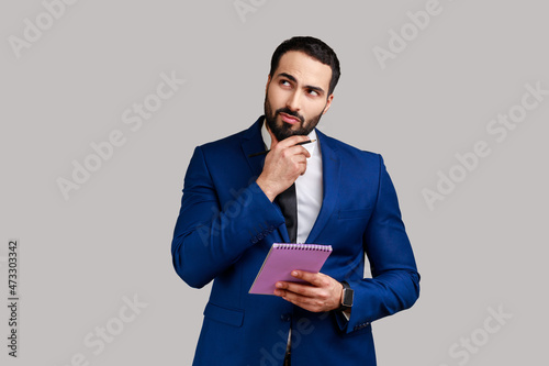 Young adult businessman standing with thoughtful expression, holding paper notebook, pondering business idea, future plans, wearing official style suit. Indoor studio shot isolated on gray background.