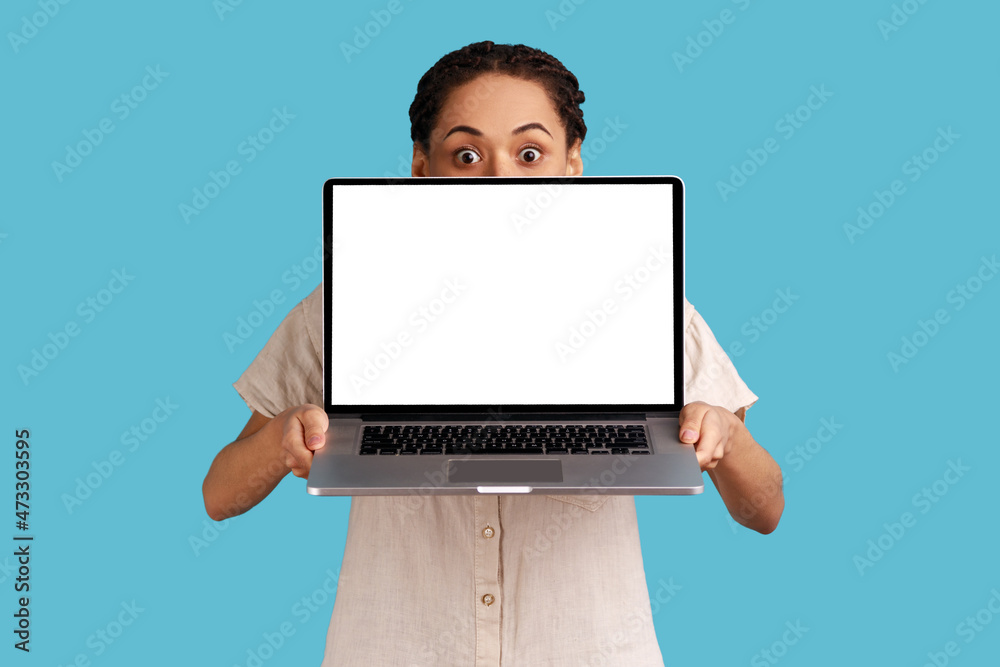 Naklejka premium Portrait of unknown woman with dreadlocks covering half of face with laptop with empty display for advertisement or promotional text, wearing shirt. Indoor studio shot isolated on blue background.