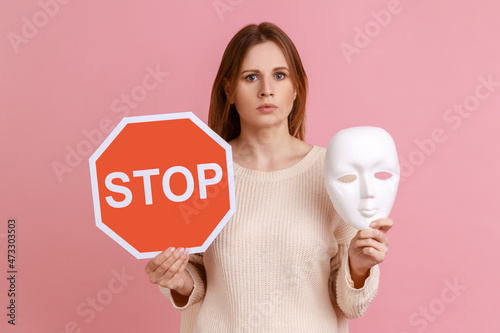 Portrait of serious blond woman holding red stop sign and white mask, looking at camera with strict expression, wearing white sweater. Indoor studio shot isolated on pink background. photo