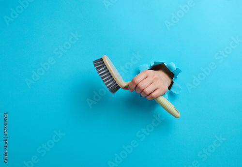 female hand holds a gray plastic brush on a blue background. A part of the body sticks out of a hole with torn edges in a paper background