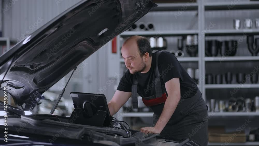 Diagnostics of the car standing at the hood in the car service. A mechanic in a close-up car service holds a tablet in his hands and presses the screen 