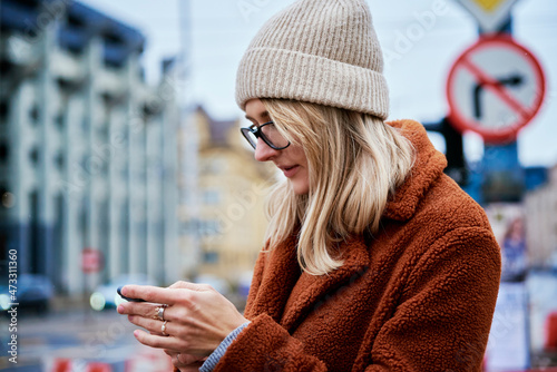 Beautiful woman in coat and eyeglasses using smartphone at city street. Online communications
