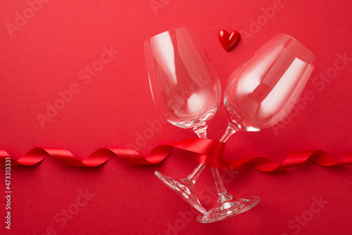 Top view photo of valentine's day decorations red curly ribbon and small heart between two wineglasses on isolated red background with copyspace