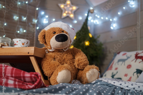 a toy teddy bear in a santa hat sits on the bed against the background of a christmas tree and a glowing garland photo