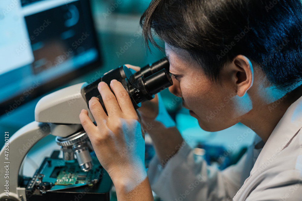 Young Asian technician looking in microscope while studying microchip characteristics in laboratory Young Asian technician looking in microscope while studying microchip characteristics in laboratory