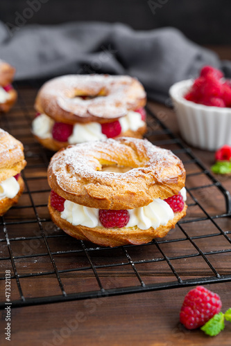 Choux pastries. Choux rings with cream of cream cheese or cottage cheese and fresh raspberries, dusted with powdered sugar on a dark wooden background.