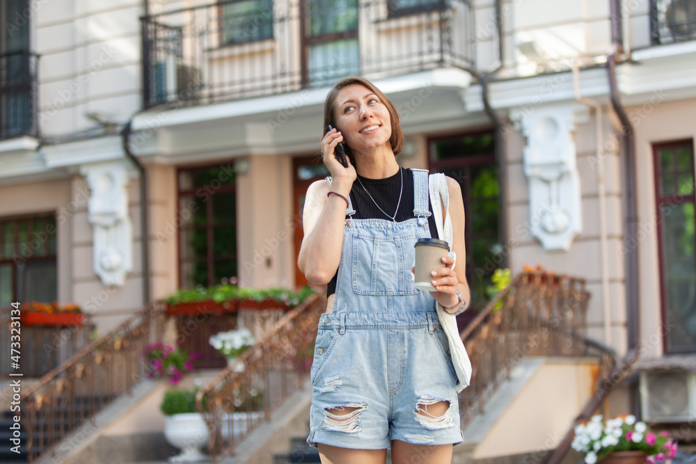 Fototapeta premium Stylish young woman in denim overalls holding cup of coffee and talking on phone in the city
