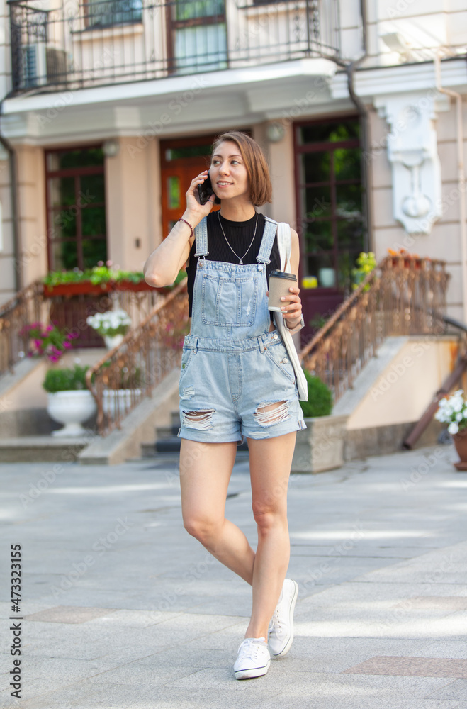 Stylish young woman in denim overalls holding cup of coffee and talking on phone in the city