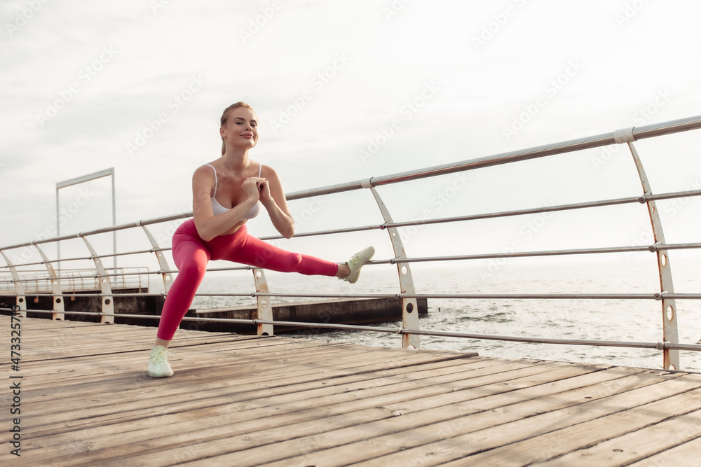 Slim fit red-haired woman in sportswear practices side lunges on the beach. Healthy lifestyle