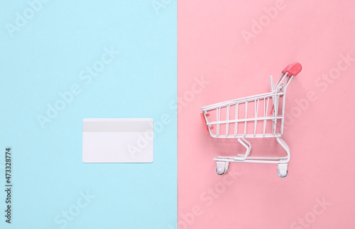 Mini shopping cart with bank card on pink blue background. Top view