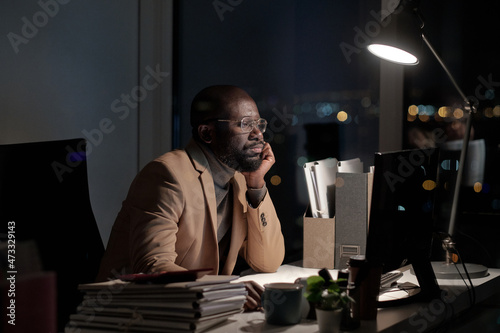 African businessman sirring by desk in front of computer monitor in office while working late at night