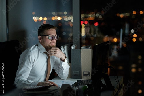 Serious broker or economist looking at computer screen while sitting by desk in dark office at night