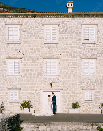 Bride and groom stand near a stone house with white blinds on the windows © Nadtochiy