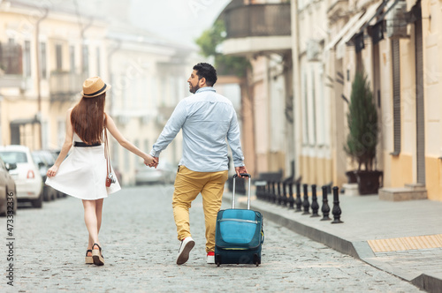 Joint, romantic trip. Loving international couple of tourists with travel luggage are walking along a European street. Spend time together.
