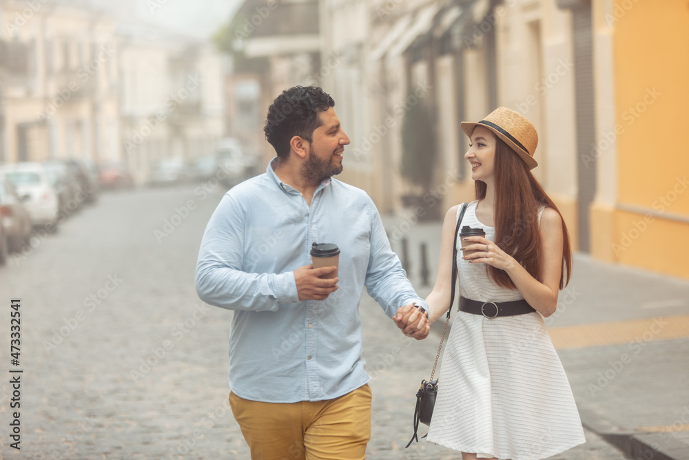 Fototapeta premium International couple in love drinking coffee walking around the city, holding hand