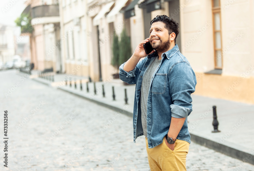 Fototapeta premium Young cheerful hispanic man in jeans jacket talking on phone on foggy european street