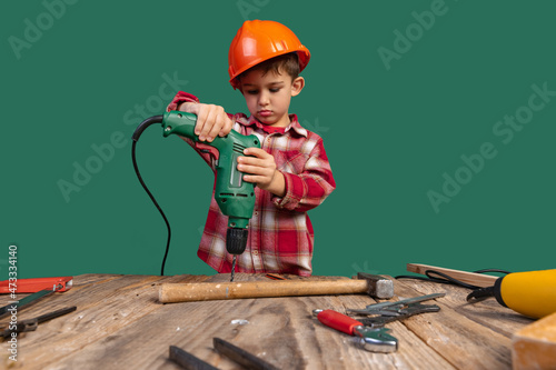 Portrait of cute little boy, kid in image of builder, designer in orange protective helmet using work tools isolated on green background photo