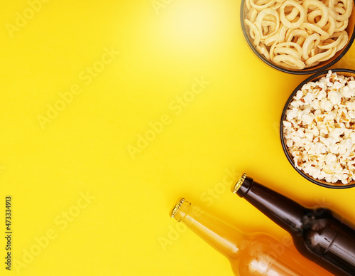 Beer bottles and bowls with snacks on yellow background. Top view